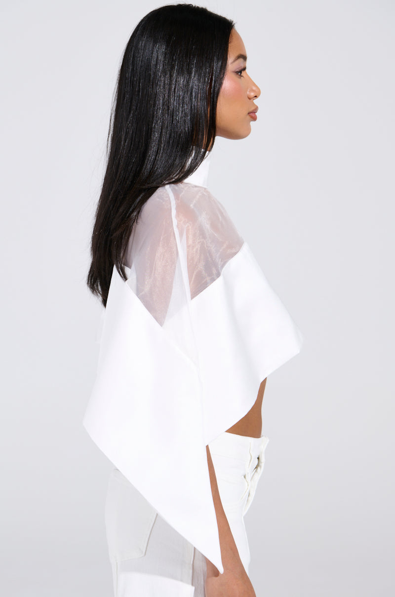 A person with long black hair stands in profile, wearing a white TALK TO ME LATER BLOUSE paired with high-waisted pants. The plain, light-colored background enhances the outfit's elegance.