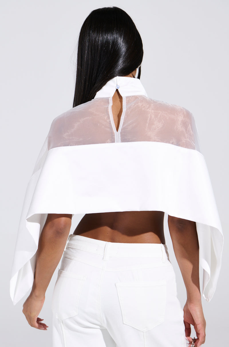 Rear view of someone with long dark hair in the TALK TO ME LATER BLOUSE, a white sheer mesh top with a high collar, cape-like silhouette, and solid bottom. It features a back keyhole opening and is paired with white pants against a plain light gray background.