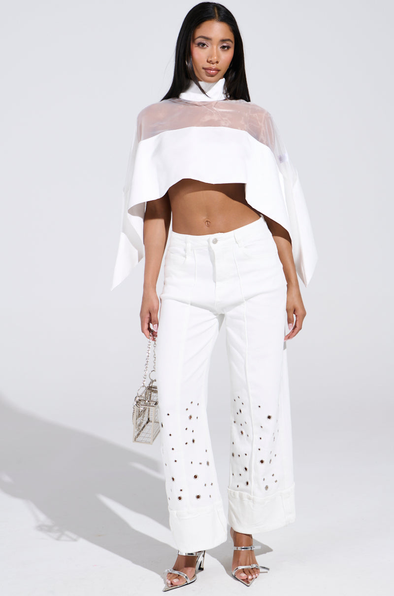 A woman stands confidently in the "Talk to Me Later Blouse," a sheer mesh top layered over a cropped white shirt and cutout white pants. She accessorizes with a small silver handbag and silver heels. A plain white backdrop emphasizes her chic and elegant silhouette.