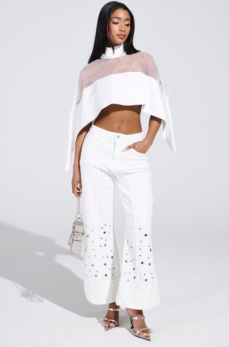 A woman stands gracefully against a plain backdrop, wearing the TALK TO ME LATER BLOUSE, paired with stylish white pants featuring cut-out details. She holds a small handbag and sports elegant high-heeled sandals.