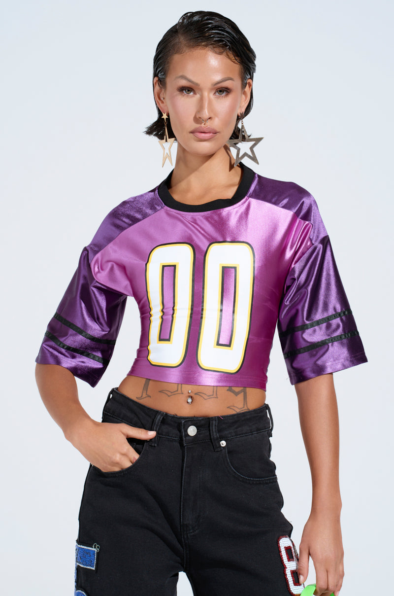 Wearing the sporty-chic DOUBLE ZERO JERSEY in a shiny purple colorway, a person confidently pairs it with black patchwork pants and star-shaped earrings. Their short, slicked-back hair contrasts the plain light-colored background.