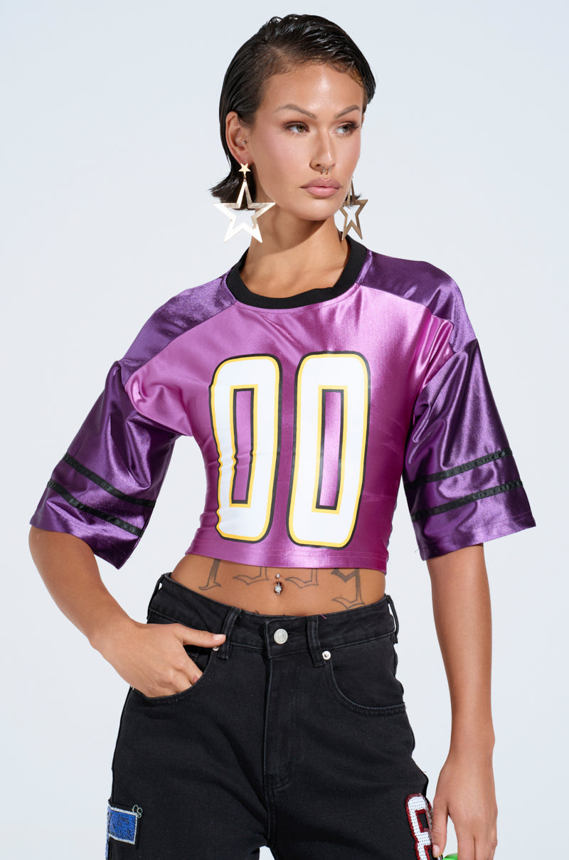 Sporting a sporty-chic look, an individual dons a purple satin crop top featuring the yellow "00" from the DOUBLE ZERO JERSEY collection. It's paired with black jeans, star-shaped earrings under slicked-back hair, and tattoos peeking above the waistline against a light gray background.