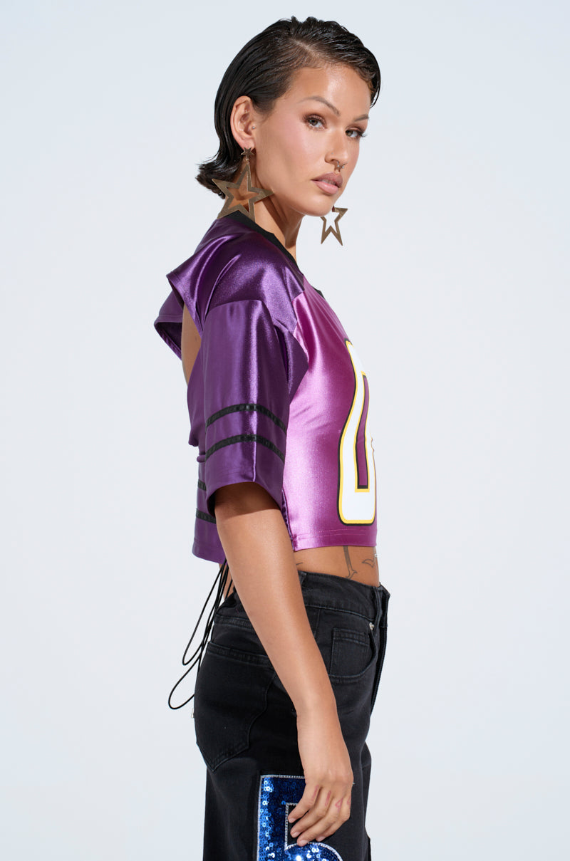A person with short hair confidently wears a DOUBLE ZERO JERSEY in purple, paired with black jeans. Star-shaped earrings and a tattoo on their exposed midriff enhance their bold style against the plain backdrop.