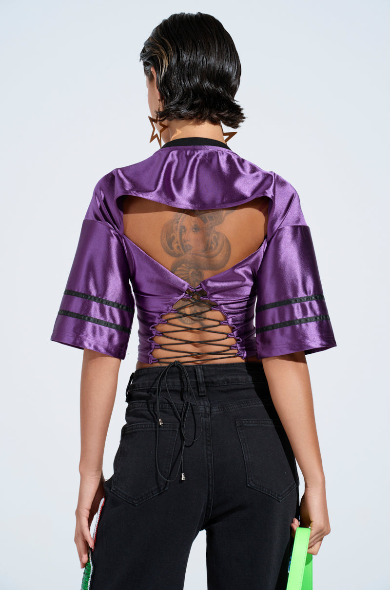 The individual with short hair turns away, flaunting a sporty-chic DOUBLE ZERO JERSEY in shiny purple with cut-out back details and black laces. They grip a green strap, complementing their black pants and star-shaped earrings, while a tattoo adorns their upper back.