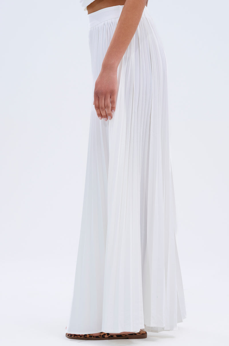 A person wearing the FLY INTO THE AIR PALAZZO PANT, a long pleated white skirt-style pant, paired with subtle leopard print flats, is shown in side profile against a plain white background.