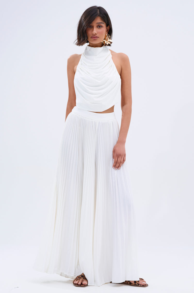 A woman with short dark hair, large statement earrings, and flat sandals stands against a plain background wearing a sleeveless white draped halter top and high-waisted, wide-leg FLY INTO THE AIR PALAZZO PANT.