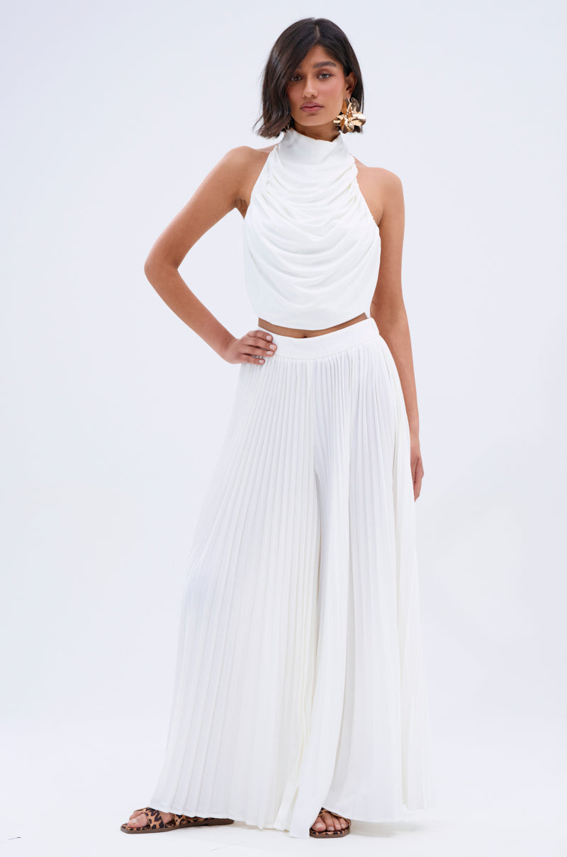 A woman poses against a white backdrop wearing the FLY INTO THE AIR PALAZZO PANT with a sleeveless white halter top, statement earrings, and leopard print sandals for a chic, airy look.
