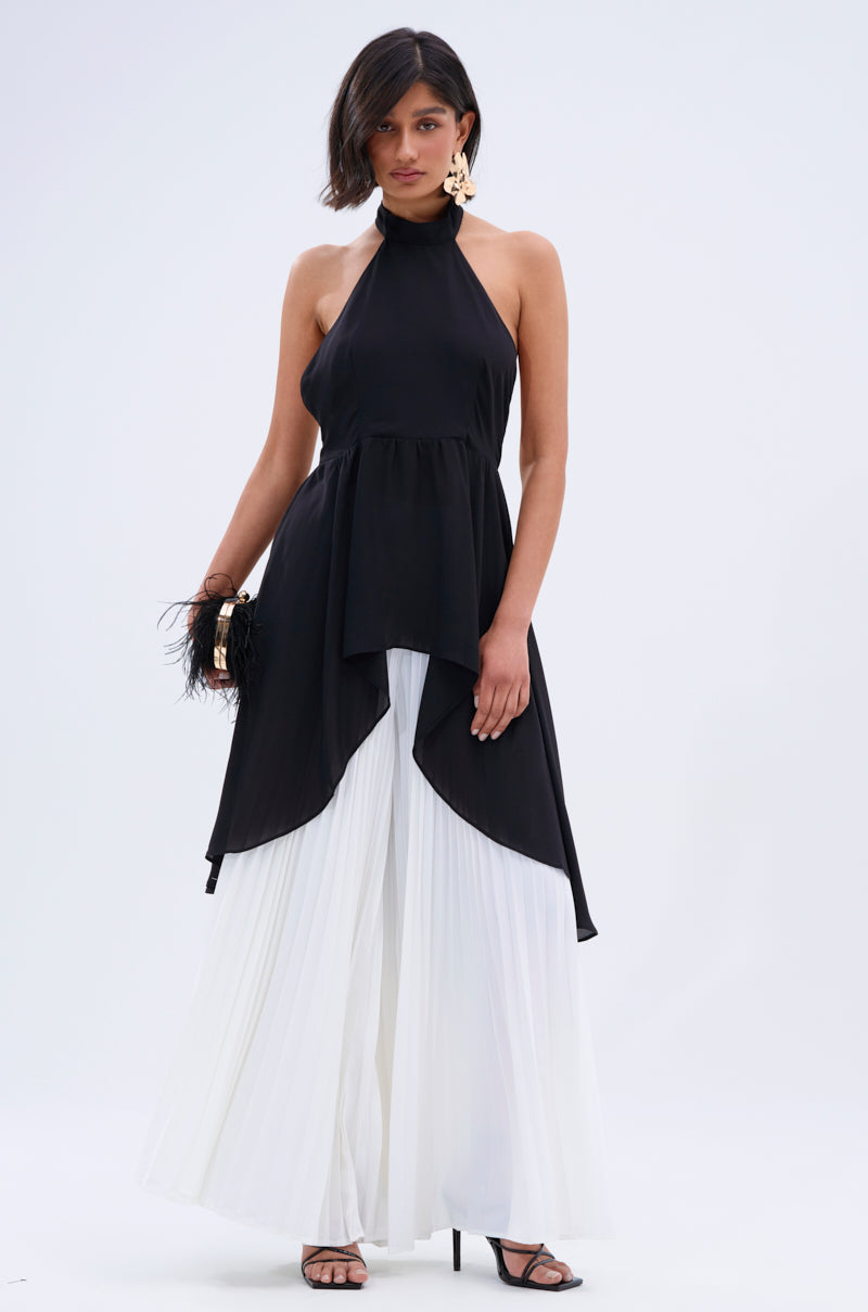 A woman in a sleeveless black halter top with an asymmetrical hem, FLY INTO THE AIR PALAZZO PANT, black heels, large gold earrings, and holding a feathered black clutch.
