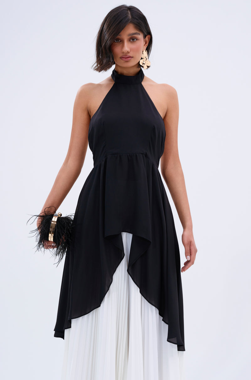 Wearing the FLY INTO THE AIR BLOUSE—a sleeveless high-neck chiffon top with an asymmetrical hem—over a white pleated skirt, she holds a black feathered clutch and sports statement earrings against a plain light backdrop.
