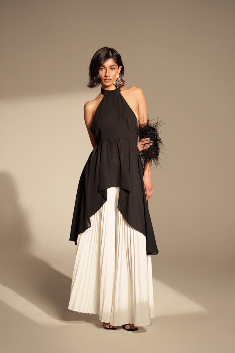 A woman with short dark hair and statement earrings wears the FLY INTO THE AIR BLOUSE—a sleeveless, high-neck top with an asymmetrical design—paired with a long white pleated skirt. She holds a black feathered accessory in a studio setting.