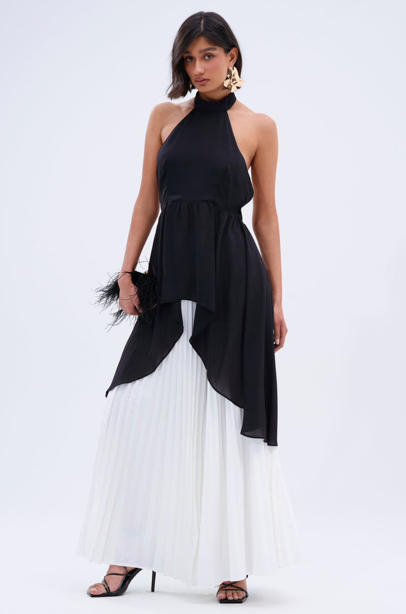 A woman wears the FLY INTO THE AIR BLOUSE, a black sleeveless high-neck top, over a long white pleated skirt. She styles it with statement earrings, chic black heels, and a black feathered clutch against a plain light background.