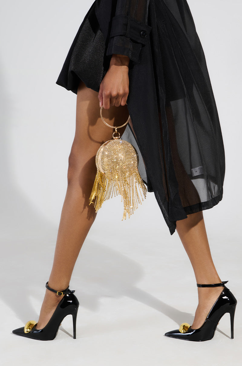 A person in a black dress with sheer layers and AZALEA WANG ESMARIA BLACK STILETTO PUMPS is walking. They are holding a gold circular handbag adorned with fringe. The plain background highlights the ensemble, while comfort cushion insoles provide effortless steps.