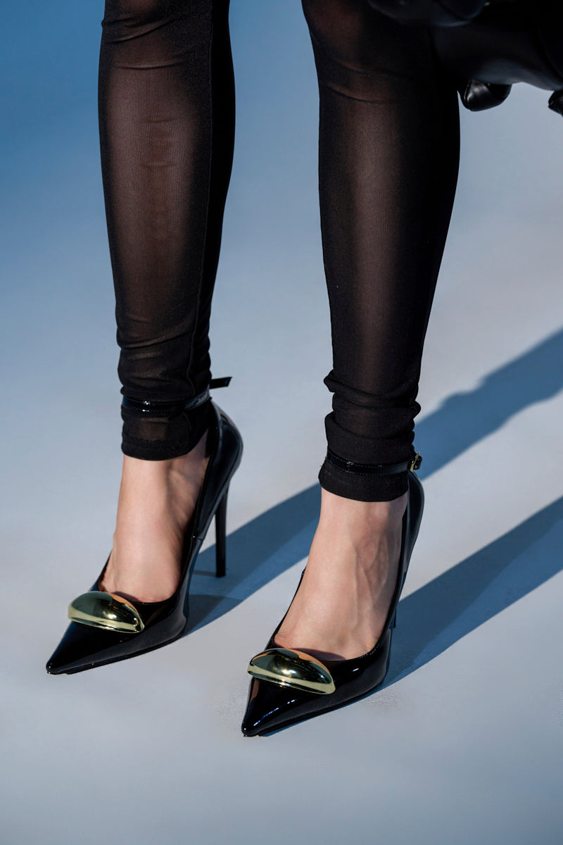 A person wears sheer black leggings and the AZALEA WANG ESMARIA BLACK STILETO PUMP, featuring a pointed toe, shiny gold accents on the front, and a comfort cushion insole.
