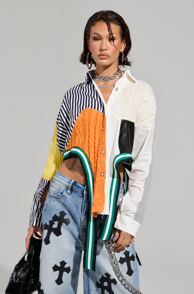 A person showcases a trendy ensemble highlighting the "Never See You Again Button Down Color Block Shirt" paired with loose jeans adorned with cross patterns. The outfit is accessorized with hoop earrings, a choker, a chain belt, and complemented by them holding a black bag. An asymmetrical cropped hemline completes the look against a neutral background.