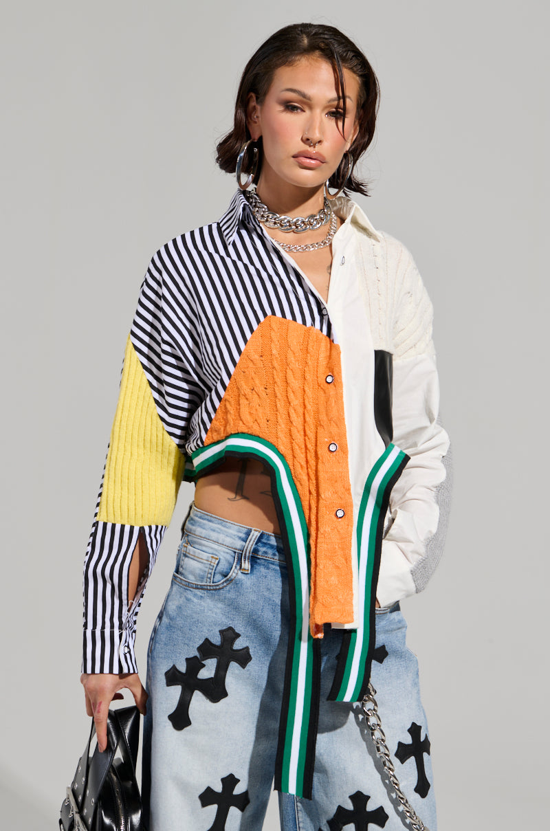 A person stands against a neutral background wearing the NEVER SEE YOU AGAIN BUTTON DOWN COLOR BLOCK SHIRT, known for its vibrant asymmetrical patterns. They pair it with light blue jeans that have cross designs, carry a black bag, and accessorize with layered necklaces and earrings.