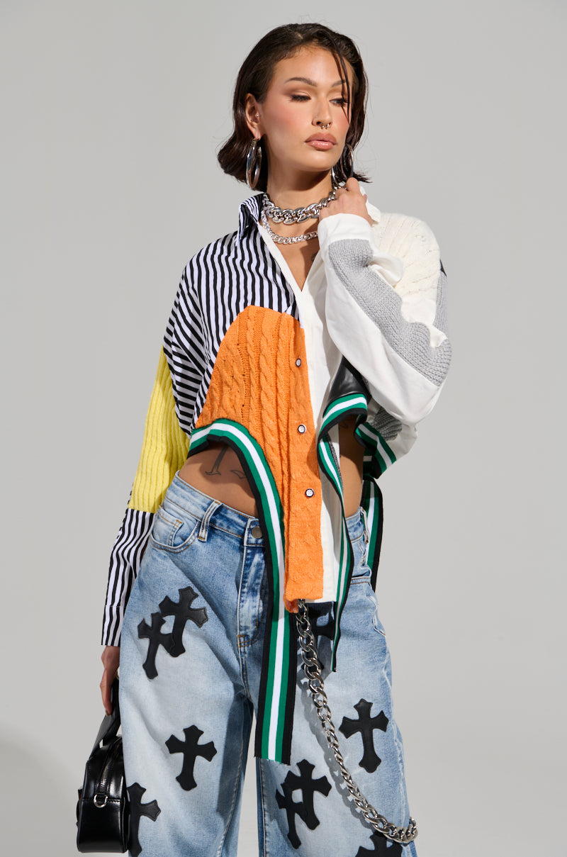 A model poses in the NEVER SEE YOU AGAIN BUTTON DOWN COLOR BLOCK SHIRT, showcasing its vibrant button-down design with stripes and cable-knit textures. She pairs it with light blue jeans adorned with black cross designs, carries a black handbag featuring a chain strap, and accessorizes with layered necklaces.