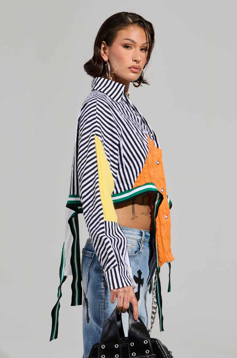 A person with short hair stands against a plain background, wearing the NEVER SEE YOU AGAIN BUTTON DOWN COLOR BLOCK SHIRT, featuring stripe print shirting with orange, yellow, and green accents. Paired with blue jeans and an asymmetrical cropped hemline, they hold a chic black studded bag.