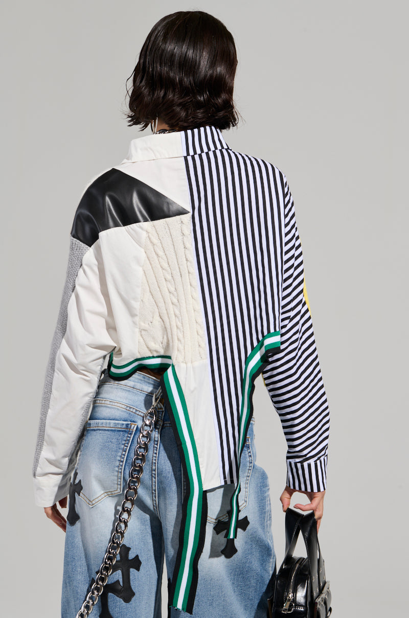 A person wearing the NEVER SEE YOU AGAIN BUTTON DOWN COLOR BLOCK SHIRT, which features a patchwork of stripe print shirting and solid colors, paired with light blue jeans adorned with black patterns. They're holding a black bag with a silver chain. The gray background subtly complements the ensemble's eclectic style.