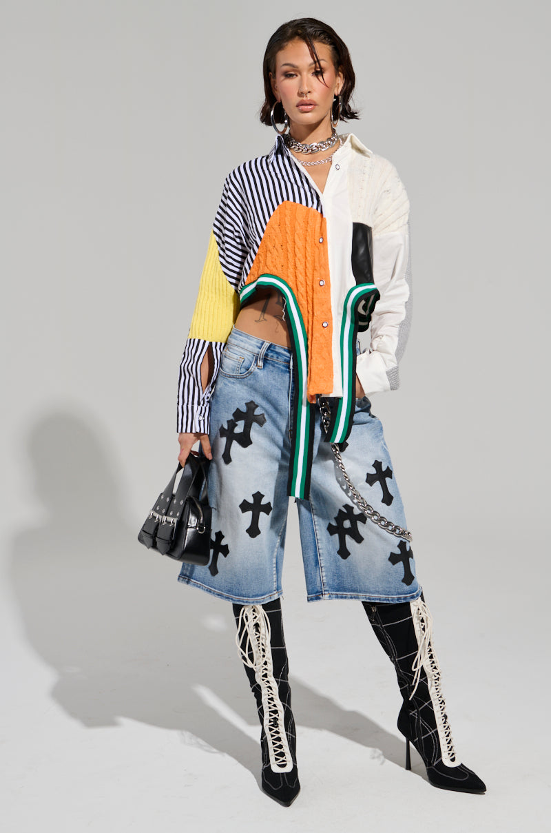 Dressed in the NEVER SEE YOU AGAIN BUTTON DOWN COLOR BLOCK SHIRT featuring a vibrant yellow, white, and black stripe print, a person stands confidently. They pair it with denim shorts adorned with black cross designs and accessorize with knee-high lace-up boots and a small black handbag.