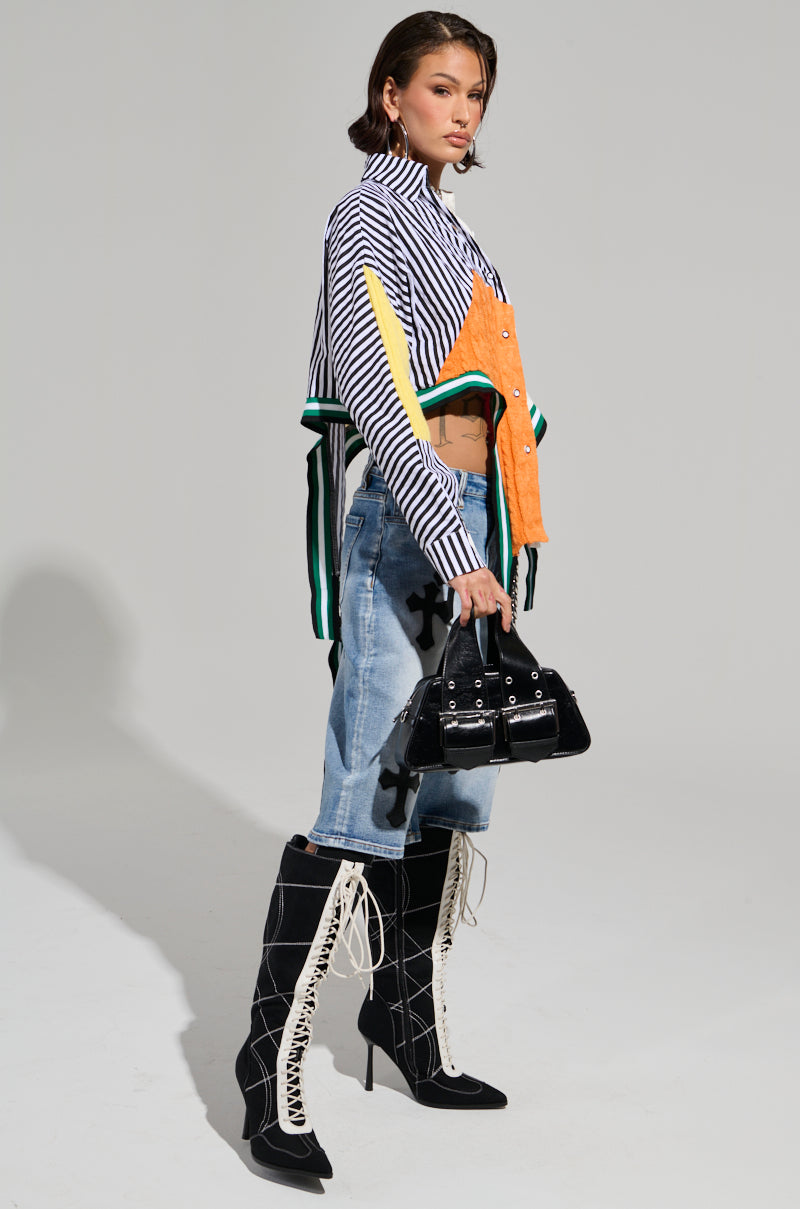 A person strikes a stylish pose in the NEVER SEE YOU AGAIN BUTTON DOWN COLOR BLOCK SHIRT with black-and-white stripes and orange accents, paired with denim shorts and knee-high black lace-up boots. They carry a small black handbag and have short hair, against a plain light gray background.