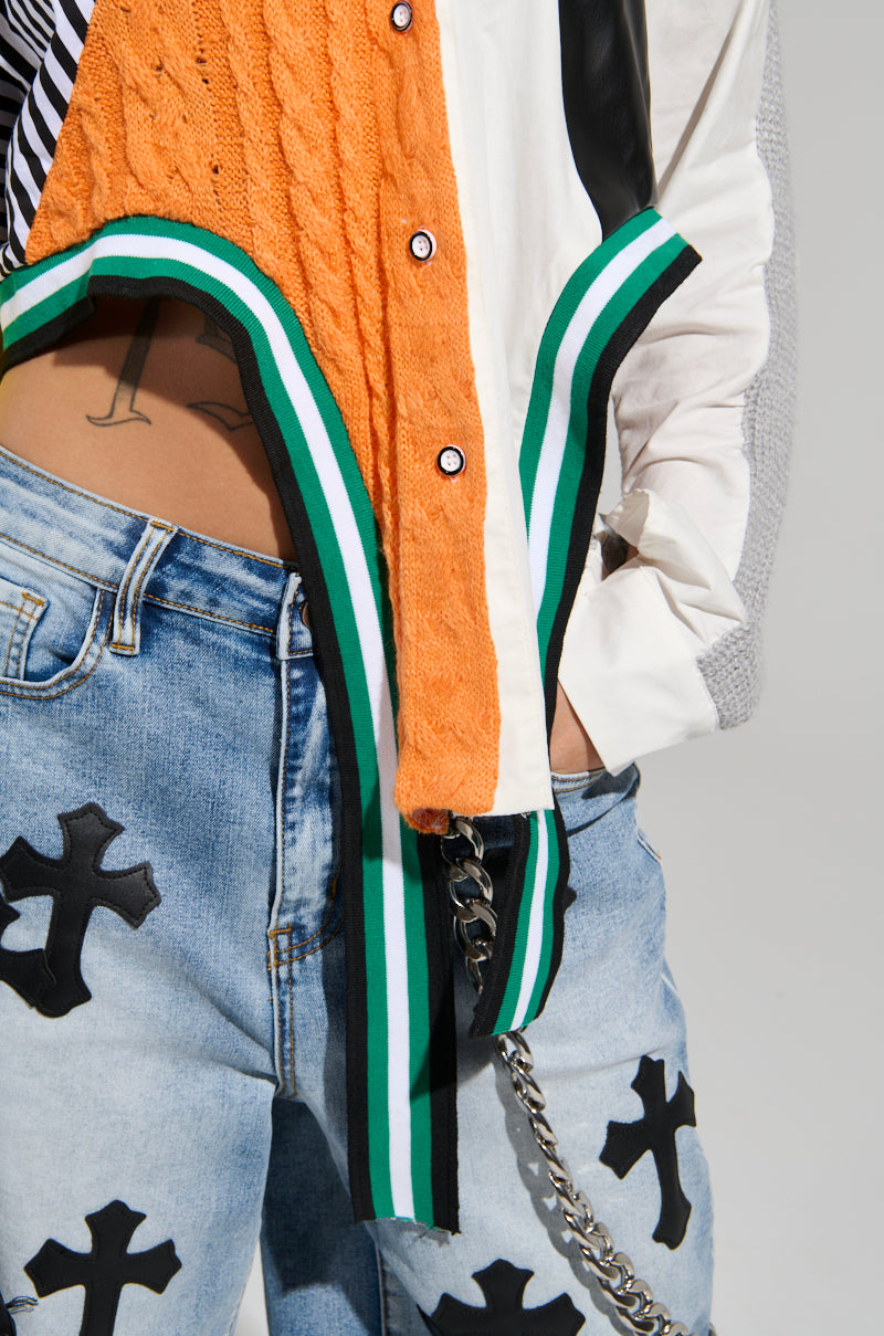A person sporting an orange sweater underneath the NEVER SEE YOU AGAIN BUTTON DOWN COLOR BLOCK SHIRT, featuring a stylish white and black design with green and white striped trim. They're also wearing blue jeans adorned with black cross patches and a chain, completing the look with an asymmetrical cropped hemline for a modern touch, while their hand rests partially in a pocket.