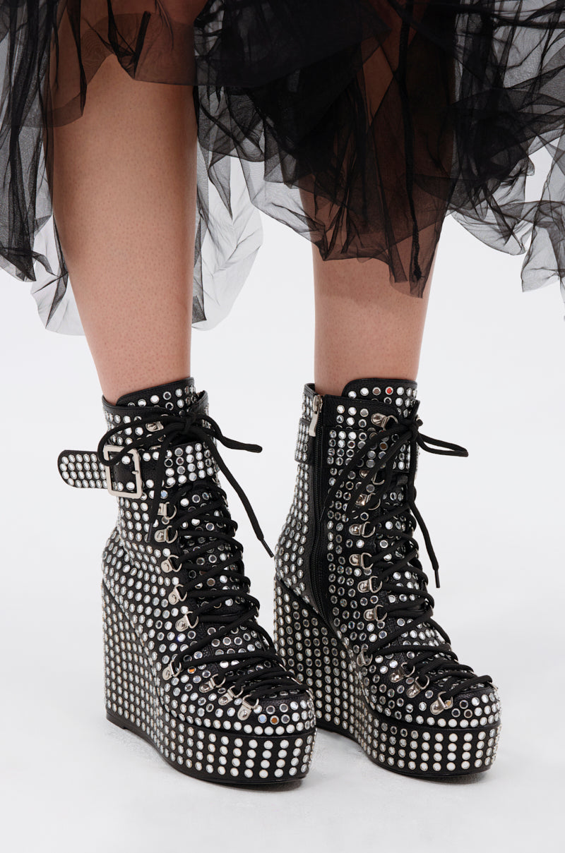 A close-up of a person wearing the AZALEA WANG FERDY BLACK PLATFORM EMBELLISHED BOOTIE, featuring silver studs, grommets, black laces, ankle straps, and paired with a black tulle skirt.