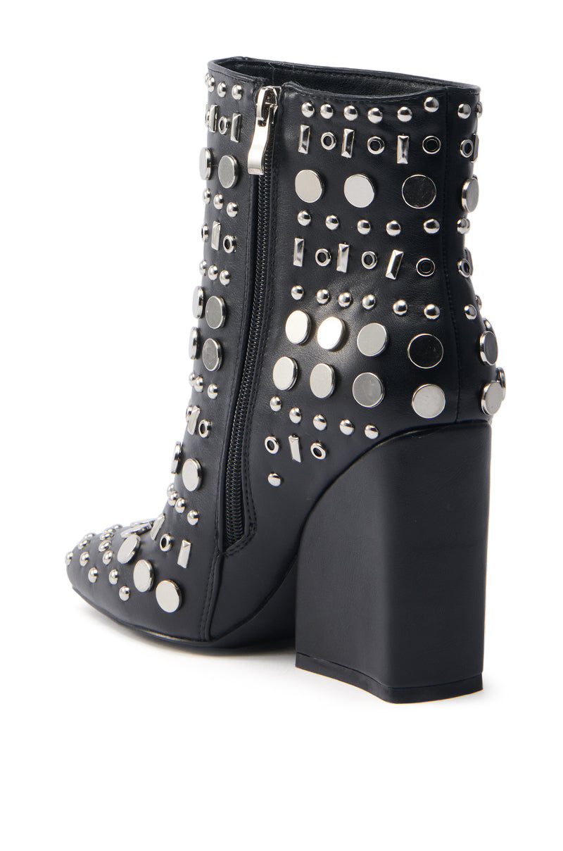 The AZALEA WANG FLETCHER BLACK SILVER METAL BOOTIE is a black ankle boot made from vegan leather, featuring a high block heel and adorned with various sizes of silver studs. It also includes a circular embellishment and a visible side zipper for easy wear.