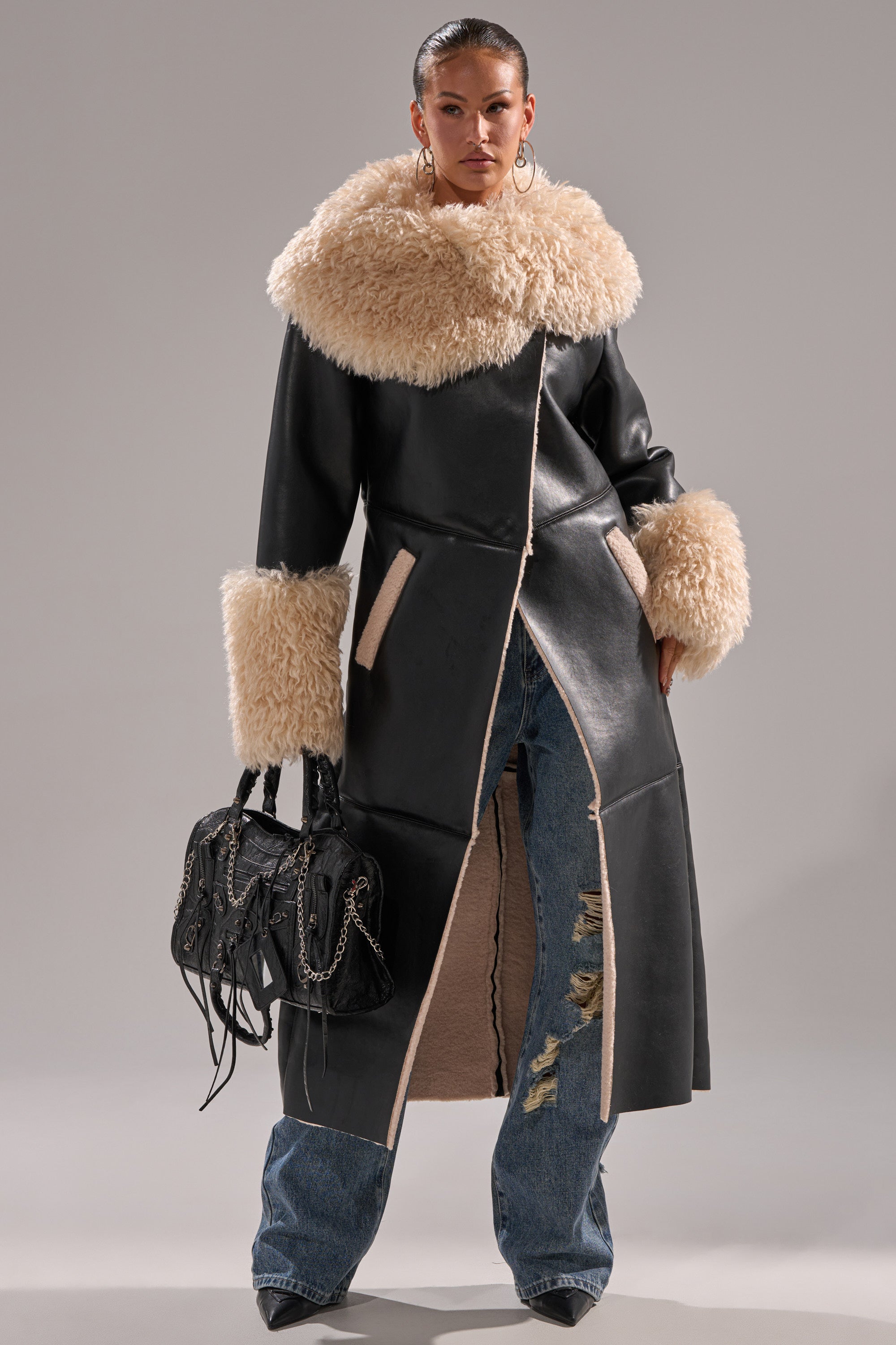 A woman models the SHE GOT IT FLUFFY PU TRENCH WITH BONDED SHERPA, featuring a long black coat with cream sherpa collar and cuffs, paired with ripped blue jeans, high heels, and a black handbag against a plain background.
