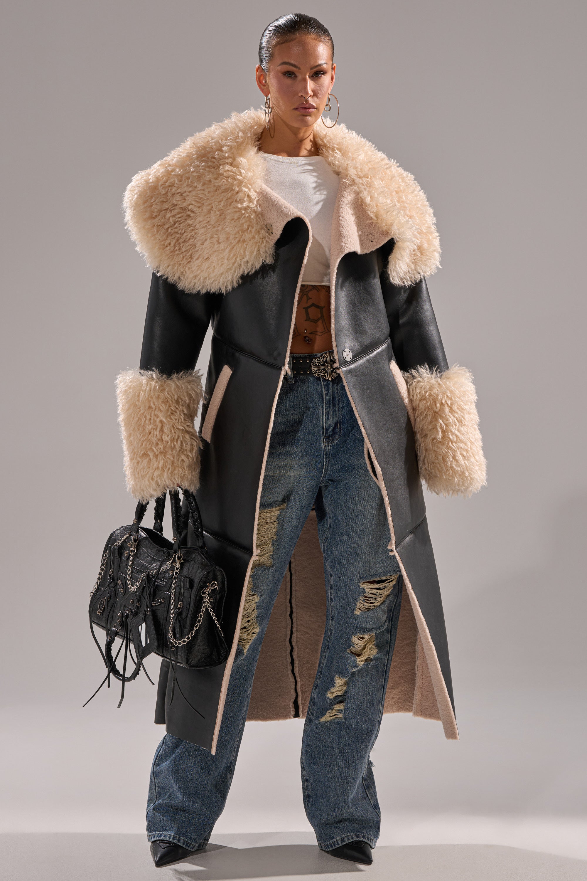 A woman wears the SHE GOT IT FLUFFY PU TRENCH WITH BONDED SHERPA, featuring a black design with a cream fur collar and cuffs, paired with a white crop top, ripped blue jeans, hoop earrings, and a black chain-detailed handbag.