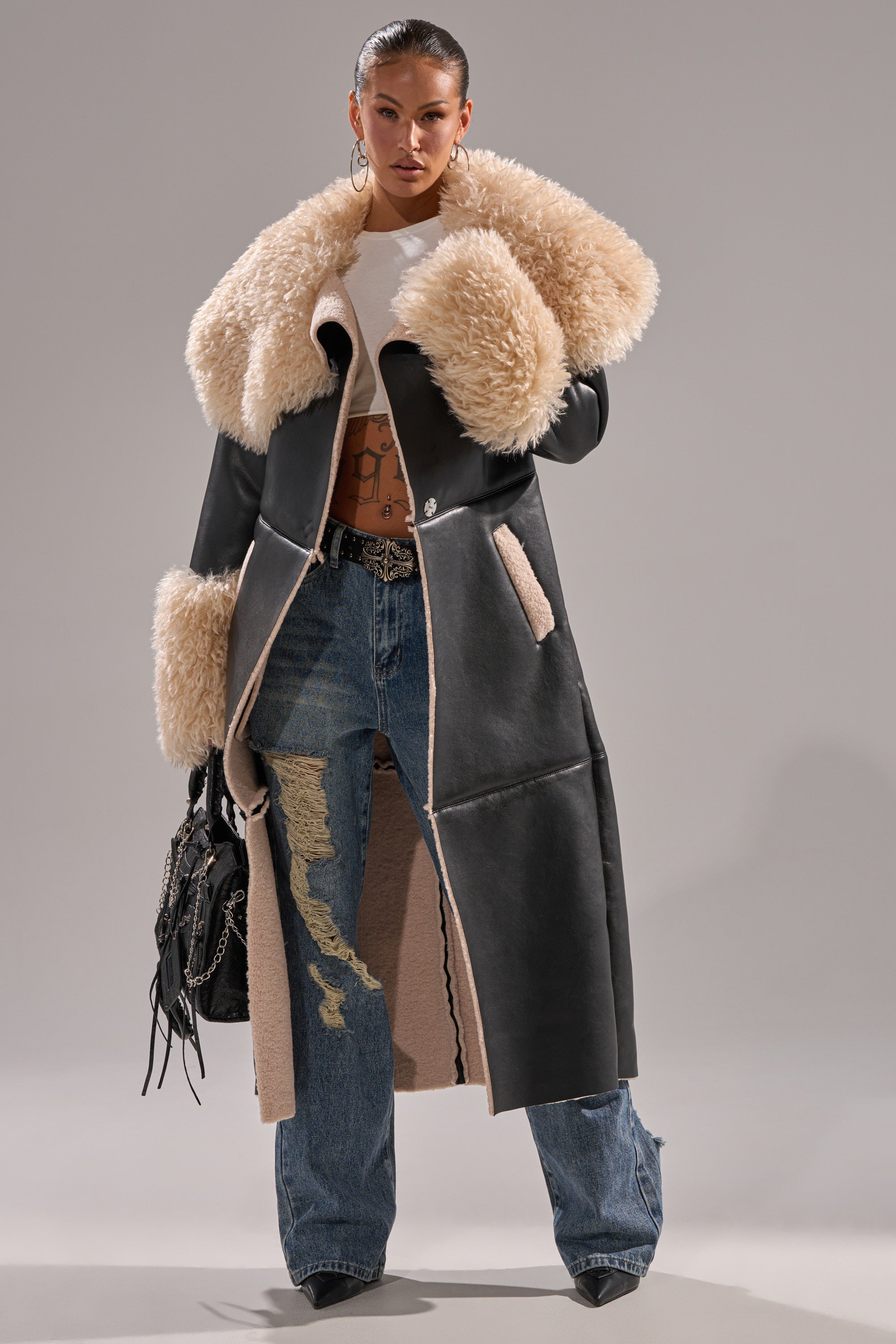 A confident woman poses in the SHE GOT IT FLUFFY PU TRENCH WITH BONDED SHERPA, featuring a long black silhouette with a cream sherpa collar and cuffs, paired with distressed blue jeans, a white crop top, hoop earrings, and a black fringe bag.
