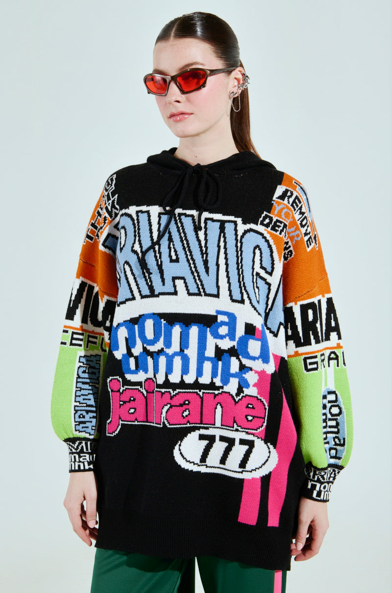 Wearing the JOYRIDE GRAPHIC HOODED SWEATER, someone showcases a vibrant colorblocking style with bold text designs like "RIAVIG" and "777." This striking look is enhanced with red-tinted sunglasses and green pants, set against a plain backdrop.