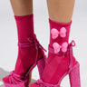 A person shows off bright pink high-heeled sandals with a chunky woven texture and ankle straps, paired with AKIRA Label's BOW DOWN SOCKS featuring matching pink bows. The stretchy knit of the socks adds a cozy touch against the plain white background.