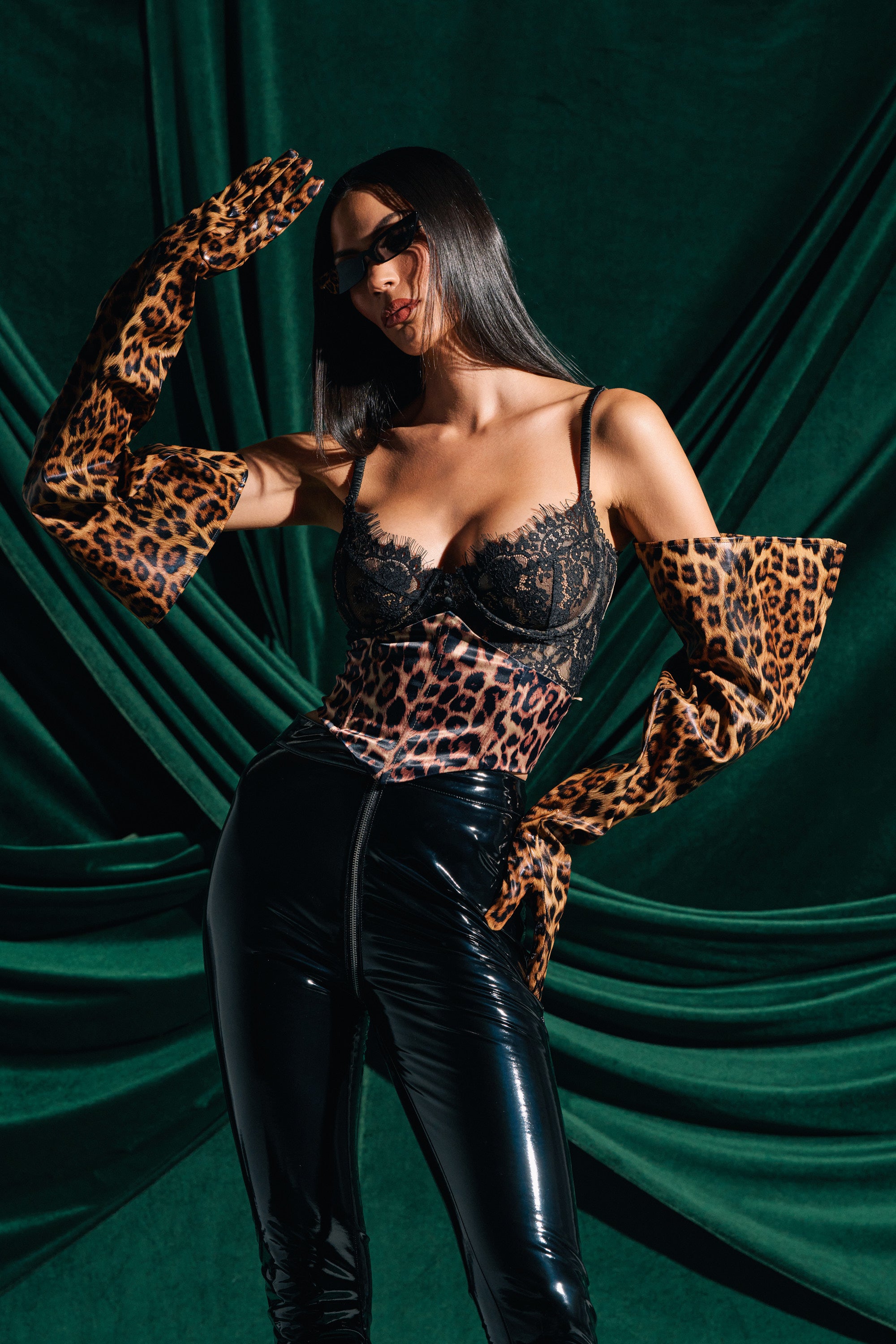 A woman exudes confidence and style in leopard print lingerie, IM STRIKING PU GLOVES, black vinyl pants, and sunglasses, posing before dark green draped fabric with straight dark hair.