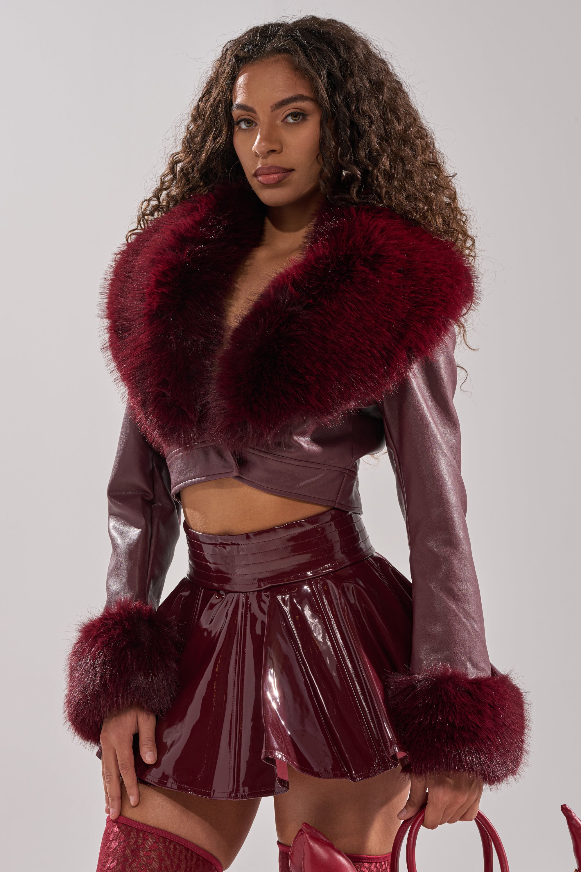 A woman wears the GISELE FUR TRIM MOTO IN BURGUNDY, a shiny cropped jacket with faux fur collar and cuffs, styled with a matching mini skirt and handbag. She has long curly hair and stands against a light background.