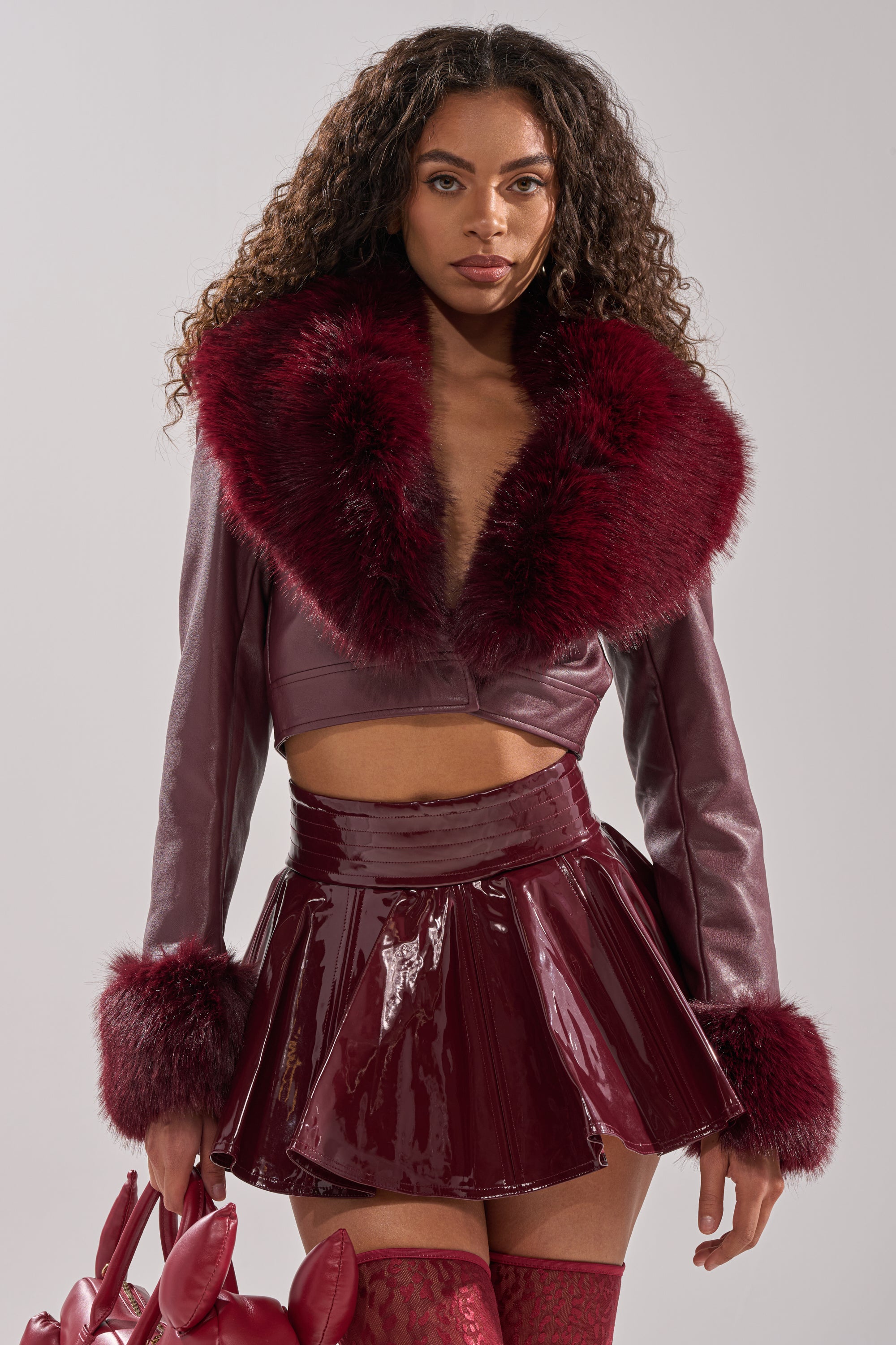 A woman wears the GISELE FUR TRIM MOTO IN BURGUNDY paired with a matching shiny mini skirt and over-the-knee stockings. She carries a red bunny ear handbag, standing against a plain background.