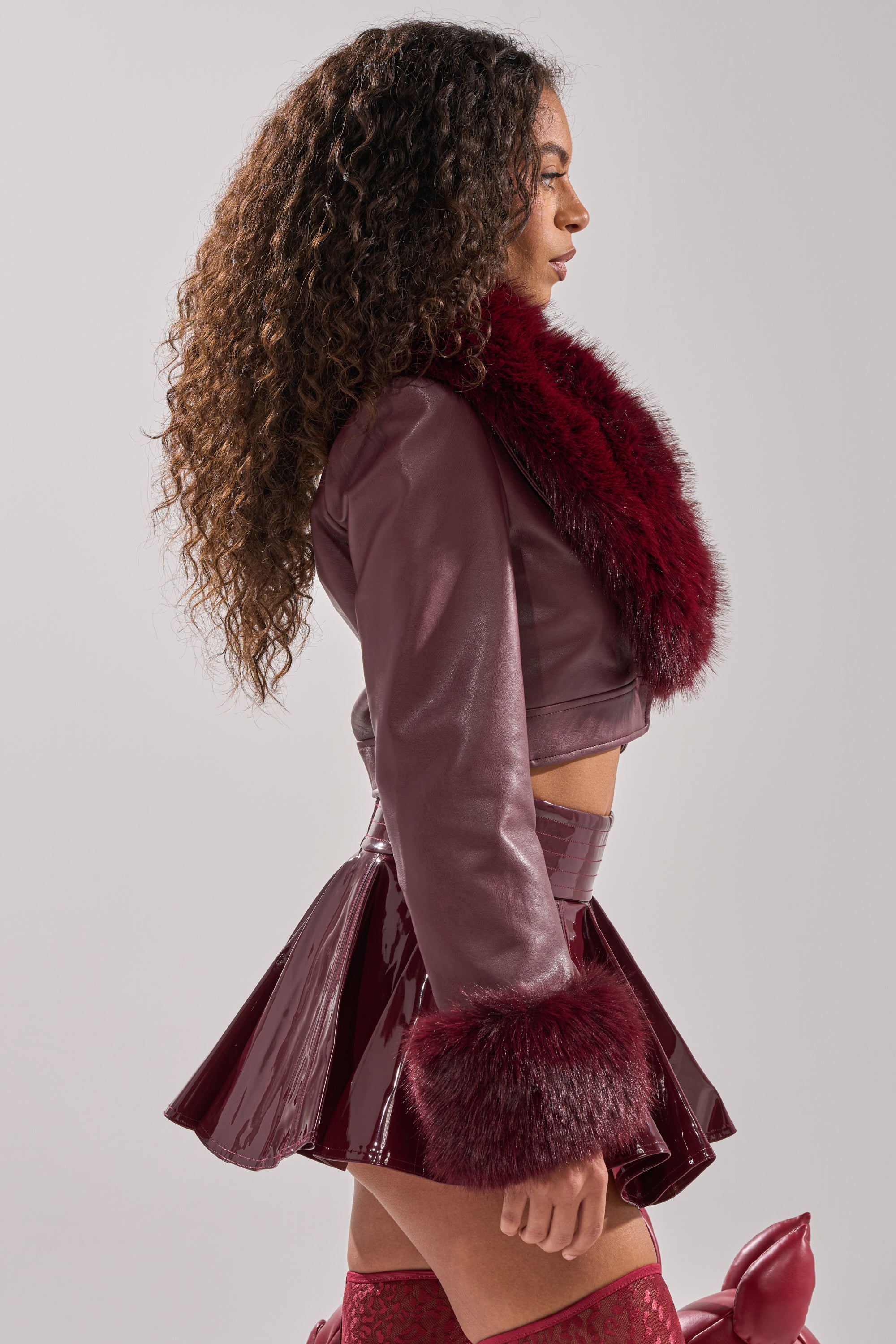 A woman with long curly hair stands in profile, wearing the GISELE FUR TRIM MOTO IN BURGUNDY and a matching pleated mini skirt. The background is plain and light-colored.
