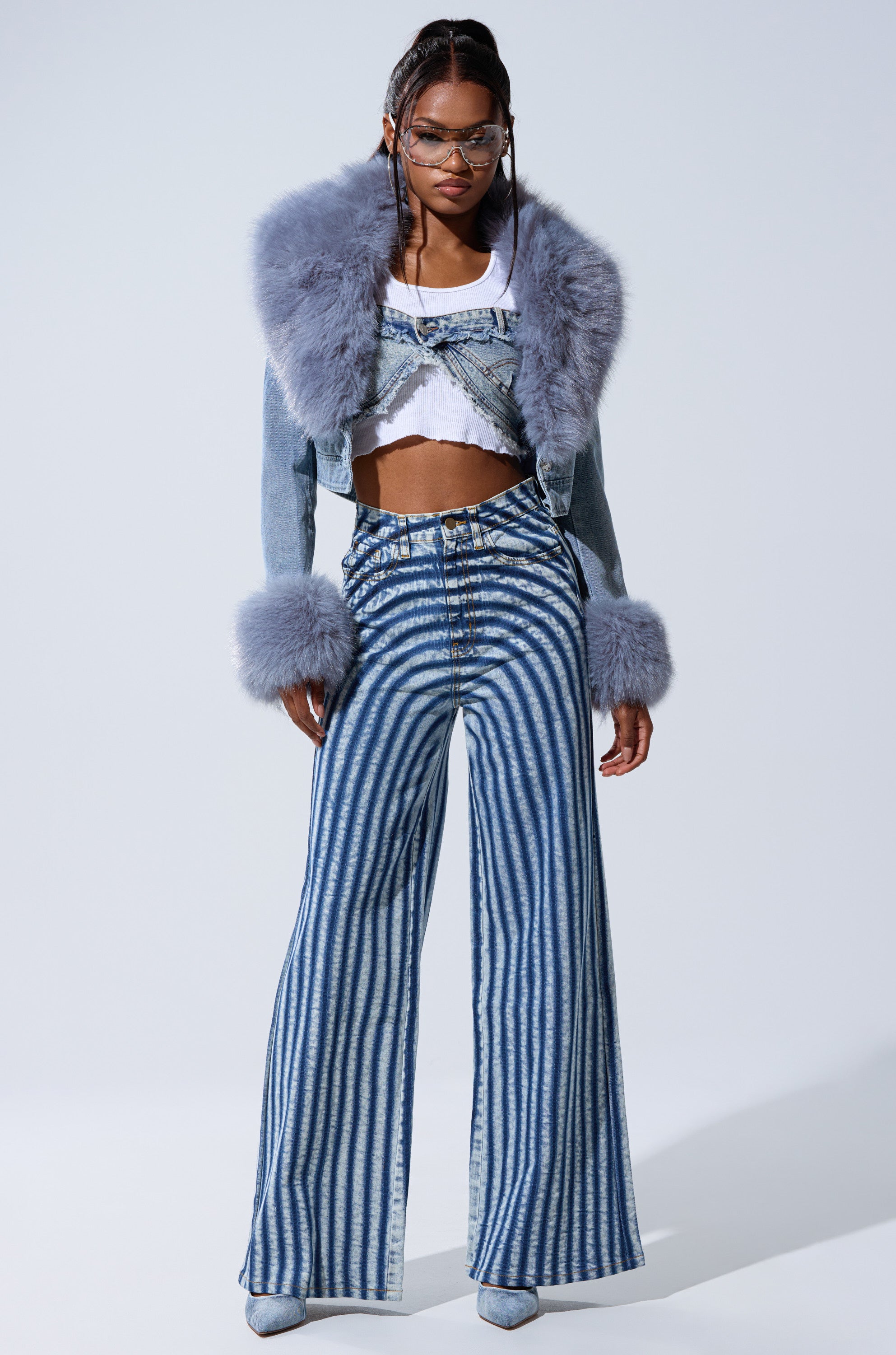 A woman poses in wide-leg, high-waisted blue and white striped jeans, a white crop top, the GISELE FUR TRIM DENIM JACKET with bold lavender cuffs and collar, blue heels, and clear glasses against a plain background.