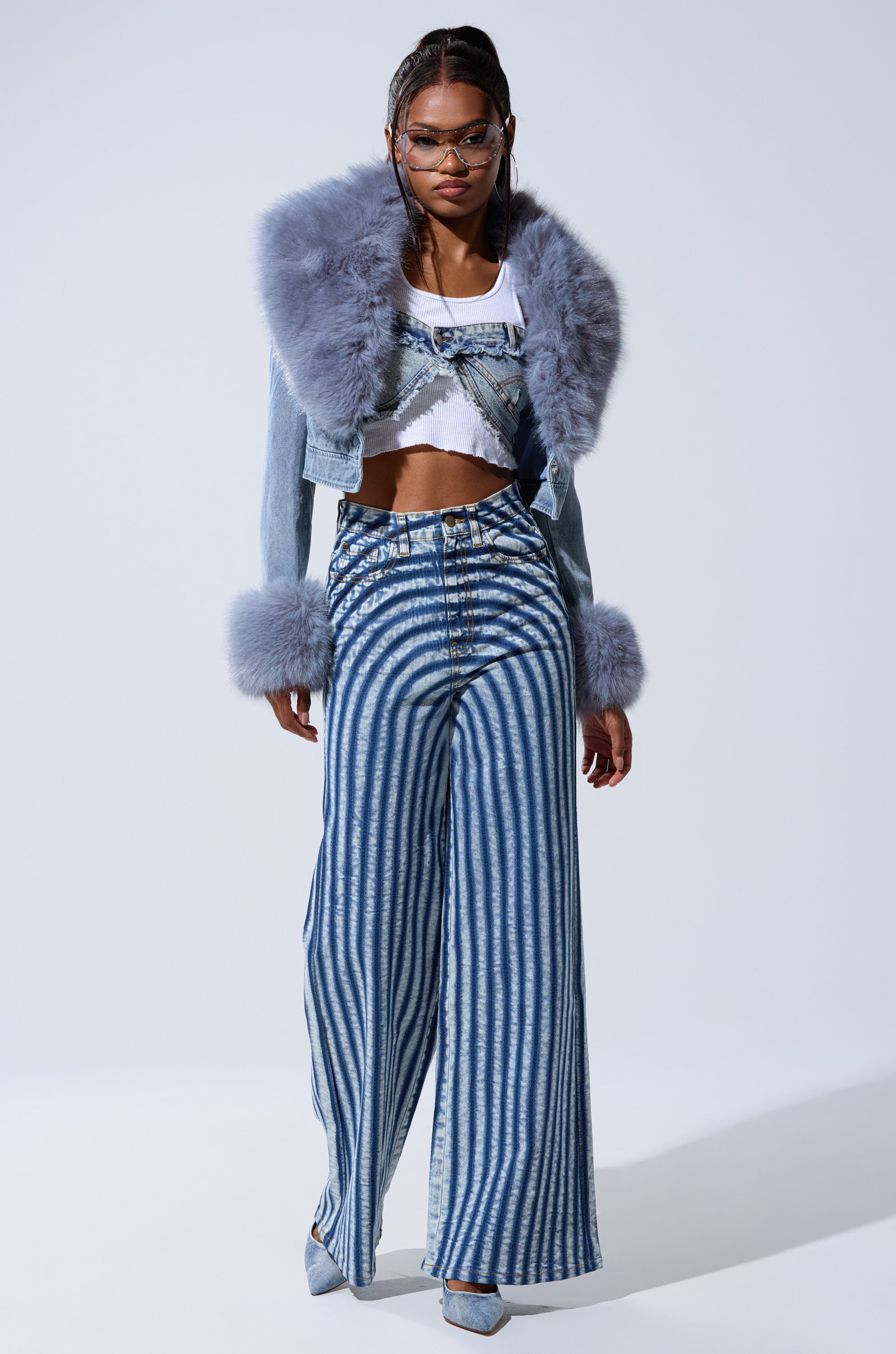 A woman models a bold look: wide-leg blue and white striped pants, a white crop top, and the GISELE FUR TRIM DENIM JACKET featuring a fluffy light purple fur collar and cuffs. She finishes the outfit with glasses and pointed heels.