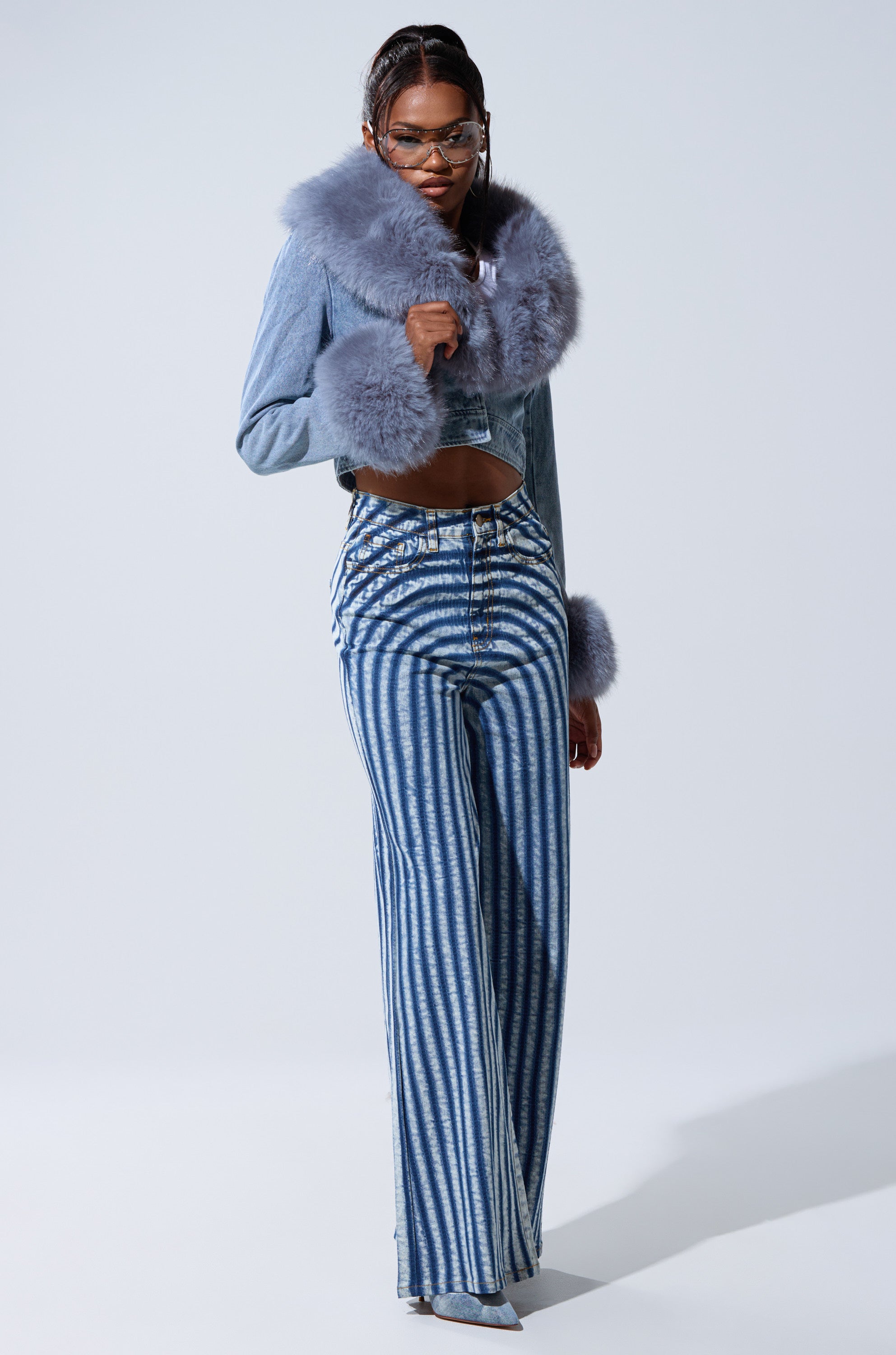A person poses confidently in the GISELE FUR TRIM DENIM JACKET, featuring large, fluffy fur trim and bold wide-leg jeans with white and blue stripes, paired with glasses and silver shoes against a light background.