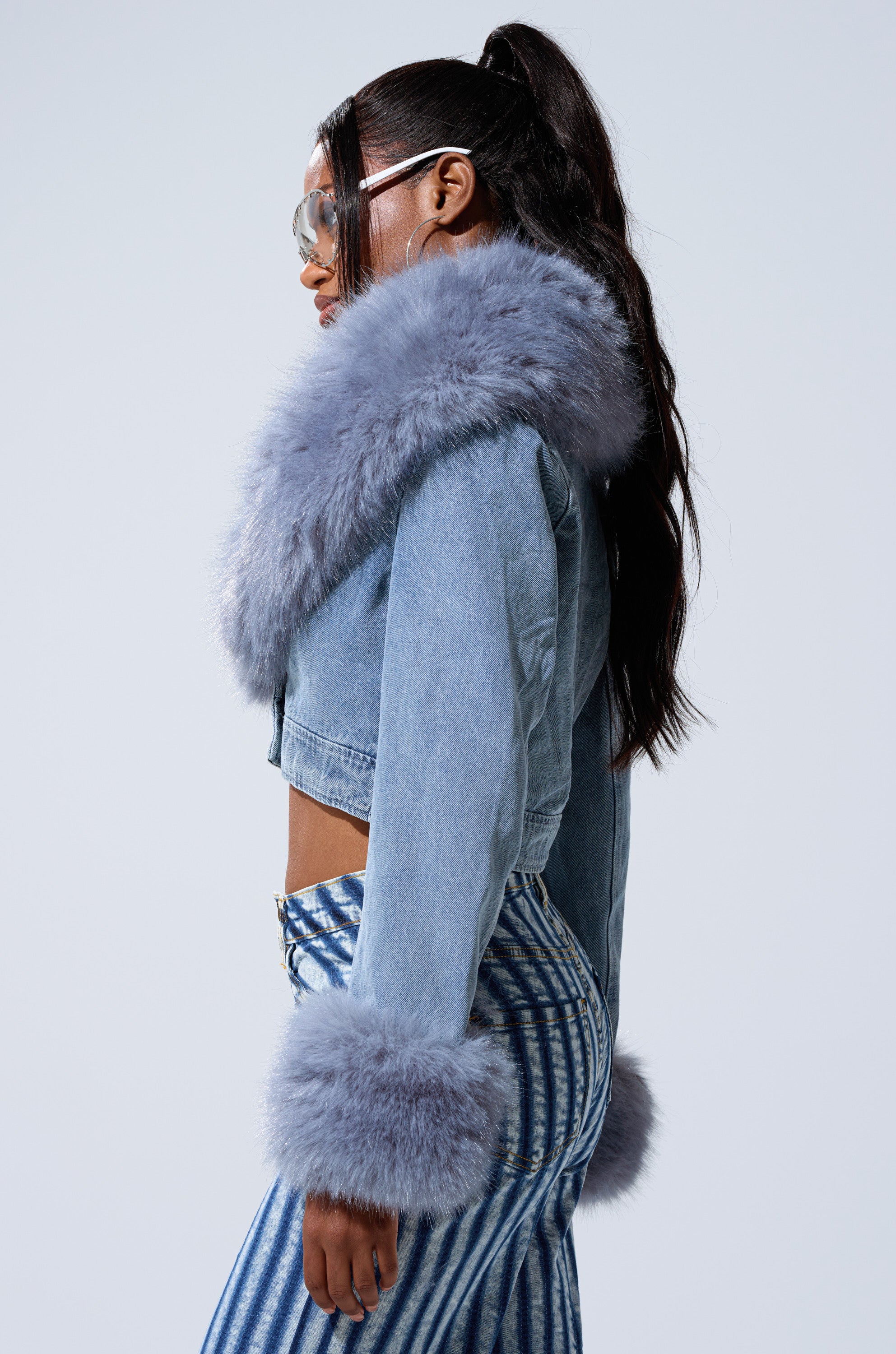 A woman in profile sports the GISELE FUR TRIM DENIM JACKET with a faux fur collar and cuffs, styled with high-waisted silver-and-blue striped pants, oversized sunglasses, and a long dark ponytail for a playful Y2K-inspired look.