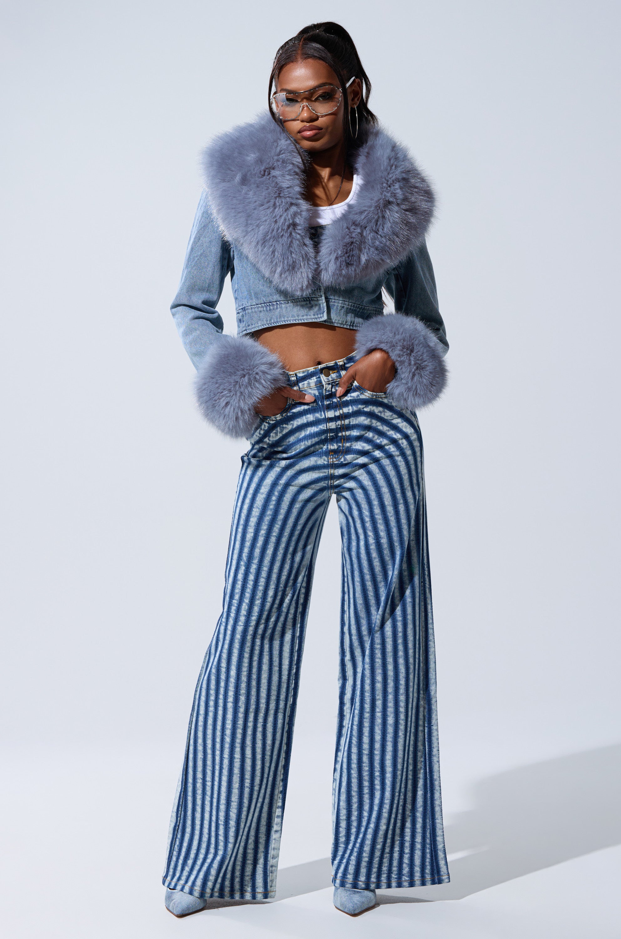 A woman models the GISELE FUR TRIM DENIM JACKET—a cropped denim piece with bold gray faux fur collar and cuffs—styled with wide-leg blue and white striped pants, pointed shoes, glasses, and hoop earrings.