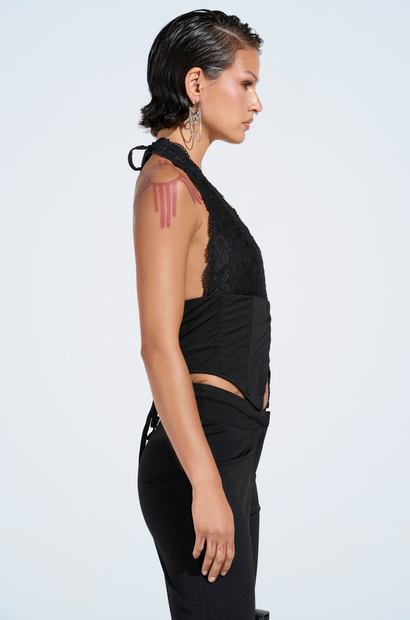 A person with slicked-back hair stands in profile, facing left, wearing the "FEELING EXTRA CORSET TOP" and black pants. A tattoo is visible on their upper arm while their hoop earrings catch the light against a plain background.