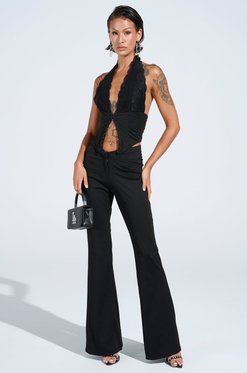 A person strikes a confident pose against a white background, wearing the FEELING EXTRA CORSET TOP in black lace with a corset neckline and black flared pants. They hold a small black bag and display tattoos on their arms.