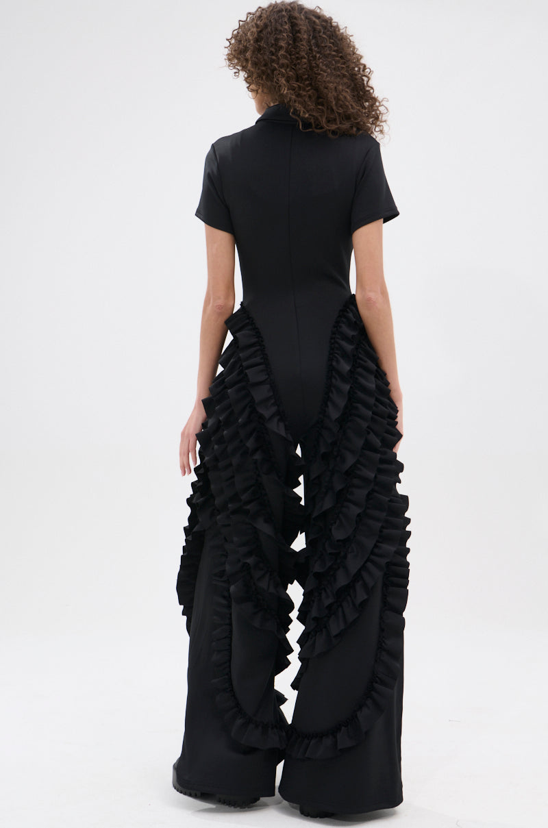 A person with curly hair models the LEAVE IT RUFFLED JUMPSUIT IN BLACK, showcasing its short sleeves and dramatic ruffled details cascading down the wide-leg silhouette. The photo is shot from behind against a plain white background.