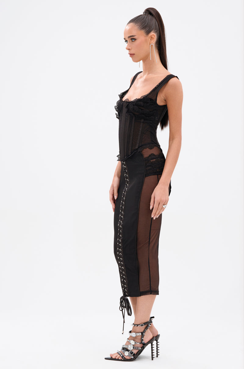 Woman standing sideways in the LET ME REMIND YOU LACE UP MESH MIDI DRESS, a corset-inspired, curve-hugging sheer black design, paired with strappy heels and long earrings against a plain white background.