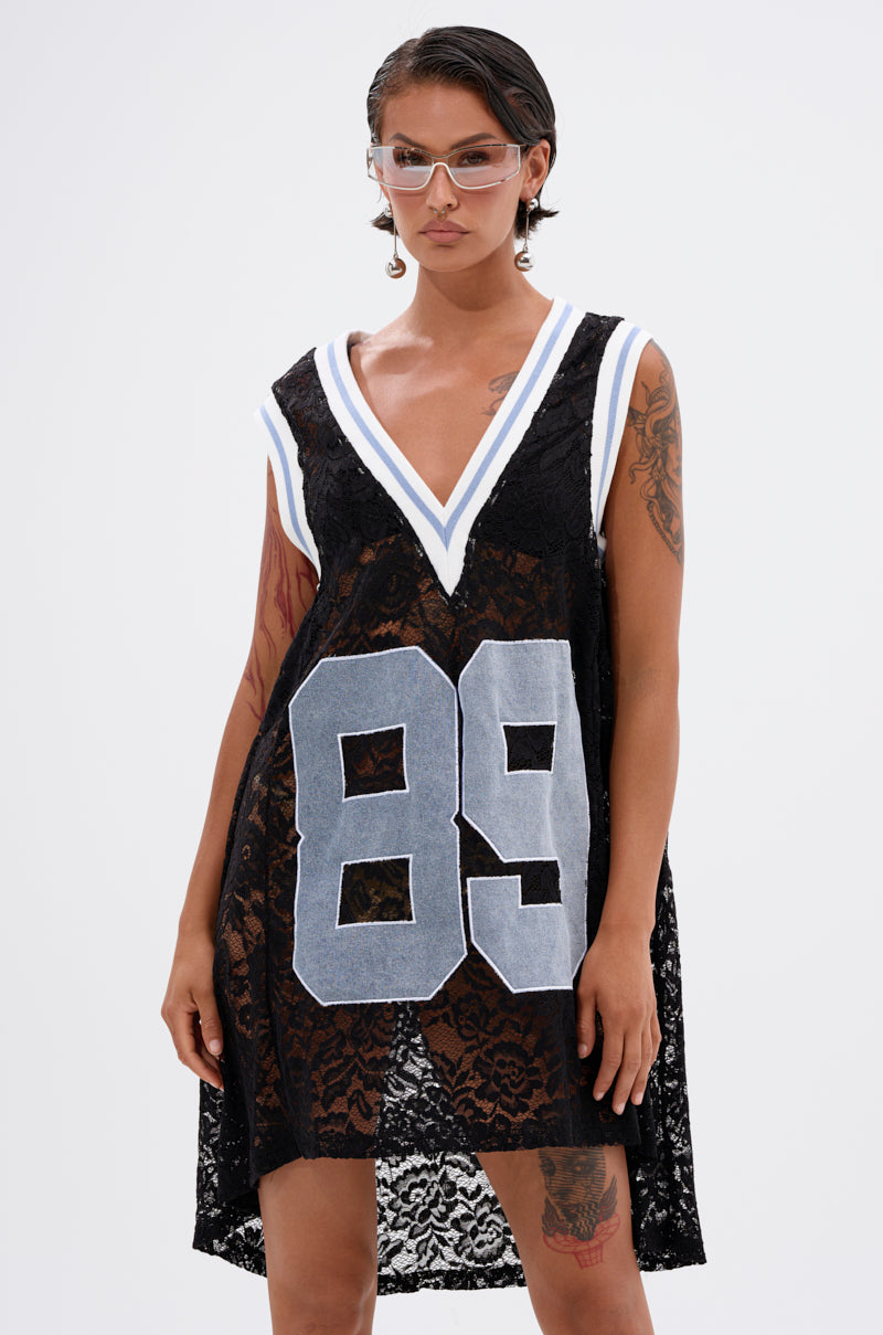 A person with short hair, tattoos, and clear glasses models the ON THE SIDELINES LACE JERSEY MINI DRESS WITH DENIM featuring a deep V-neck and bold "89" on the front. The background is plain white.