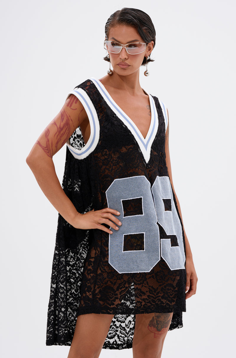 A person models the ON THE SIDELINES LACE JERSEY MINI DRESS WITH DENIM—featuring floral lace, bold white and gray "89" numbers—along with clear sunglasses and visible tattoos, against a plain white background.