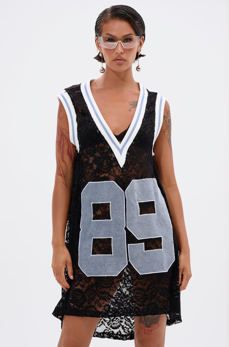 A person wears the ON THE SIDELINES LACE JERSEY MINI DRESS WITH DENIM—black lace with a deep V-neck, white and blue trim, and bold gray "89"—confidently posing against a plain white background.