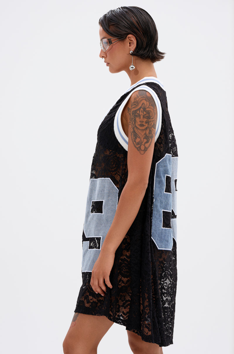 A person with short dark hair, sunglasses, a dangling earring, and arm tattoos models the ON THE SIDELINES LACE JERSEY MINI DRESS WITH DENIM, featuring a sports jersey-inspired style and the number 98 on the back. Side profile, plain background.