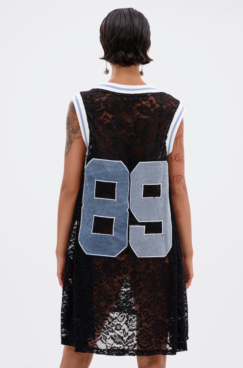 A person with short dark hair and tattooed arms faces away, wearing the ON THE SIDELINES Lace Jersey Mini Dress with Denim, featuring a sleeveless black lace design and a large light blue "89" on the back. Background is plain white.